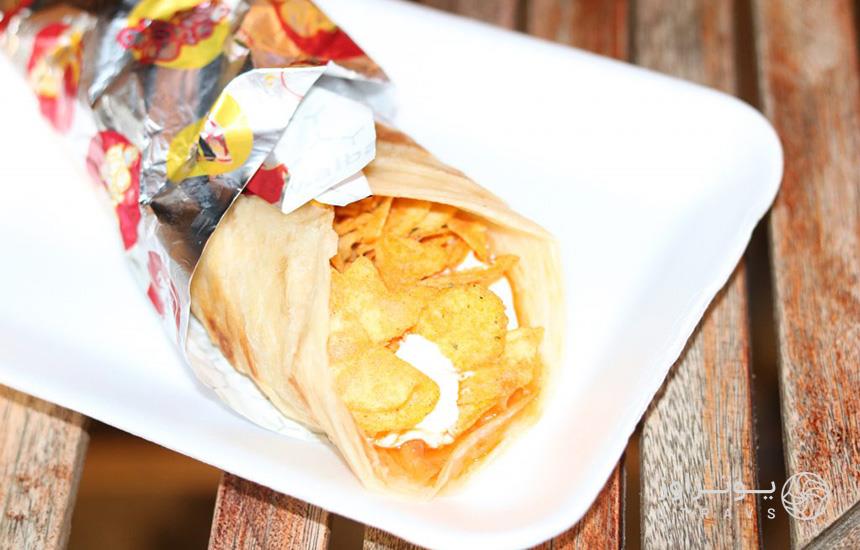 تصویری از ساندویچ رول چیپس عمان که در کاغذ زرورقی پیچیده شده‌ و داخل بشقابی سفیدرنگ قرار دارد.